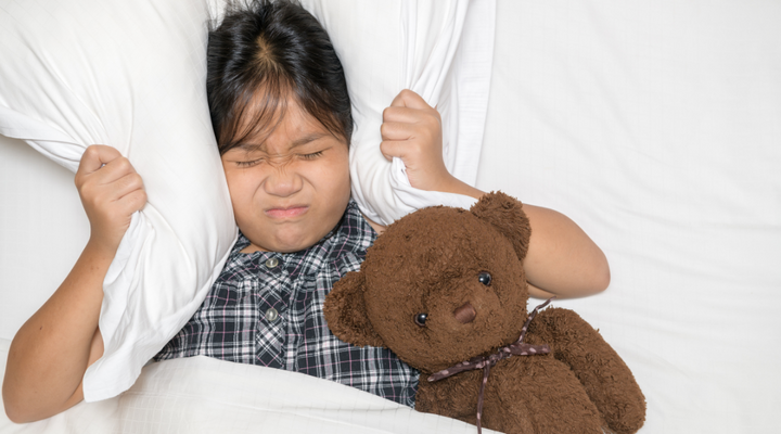 Little girl in bed covering ears with pillow to block out too loud noise and holding teddy bear 