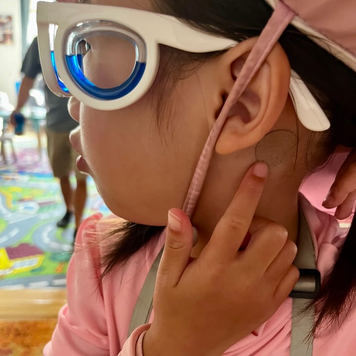 Little girl wearing anti-nausea glasses and showing anti-motion sickness patch behind her ear